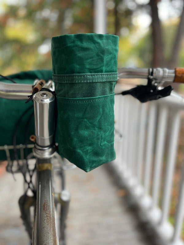 A dark green fabric bicycle handlebar bag mounted on silver bicycle handlebars in an outdoor setting with a blurred railing and trees in the background.