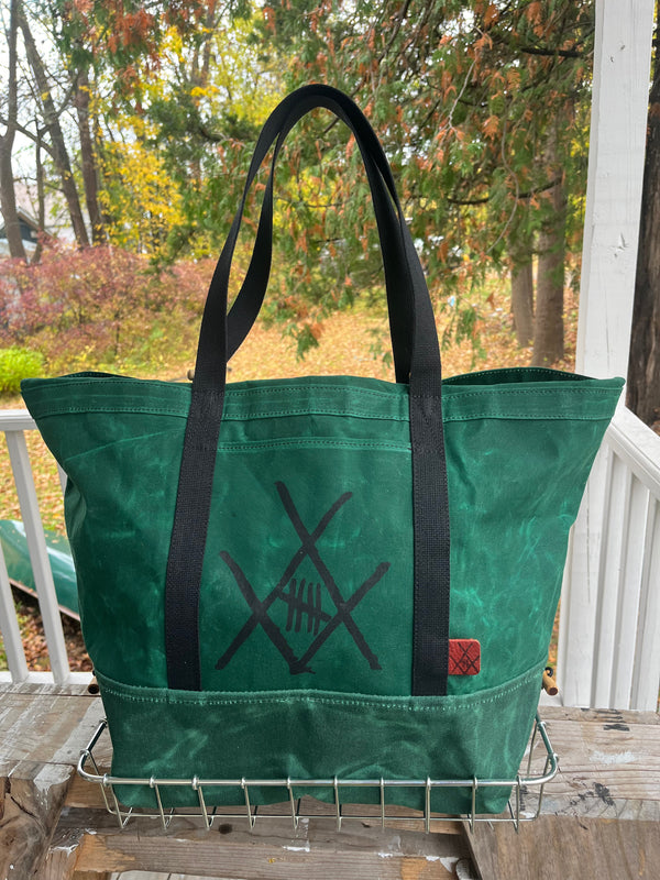 A green canvas tote bag with black handles and a black crossed-knife style graphic on the front, sitting on a metal wire rack on a porch with an outdoor autumn garden background.