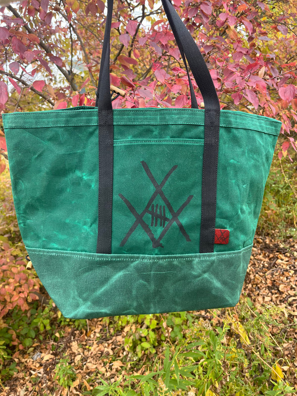 A green canvas tote bag with black handles and a black geometric cross design on the front, hanging outdoors against a background of pink and green autumn foliage.