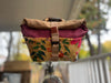 A bicycle front bag with a tan canvas top, burgundy fabric lining, and a mustard yellow floral patterned lower section, mounted on a bicycle handlebar with a blurred outdoor background.