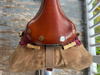 A close-up of a Brooks bicycle saddle with a brown leather top, brass hardware, and a tan canvas bag attached with a strap, photographed outdoors against weathered wooden boards.