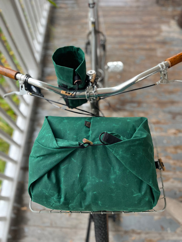 A dark green bicycle basket bag filled with a matching green fabric cover, mounted on a bicycle with wooden handlebar grips, photographed on a wooden boardwalk.