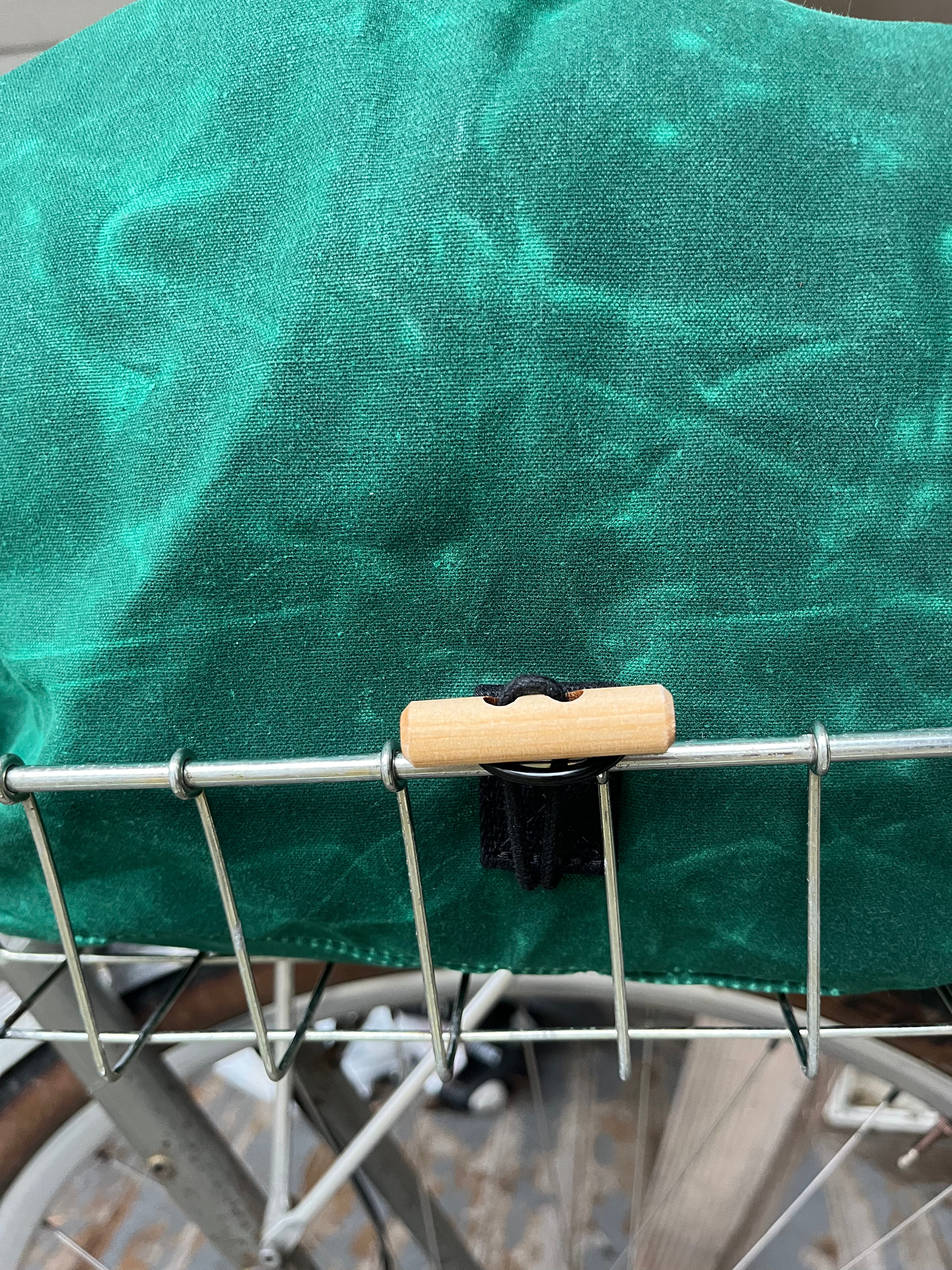 A close-up of a green bicycle pannier cover secured with a black clip and light wood handle on a metal bicycle rack.