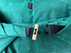 Close-up of a teal-green fabric bag or garment with a black strap and a light natural wood toggle closure, plus a small black fabric tag near the top.