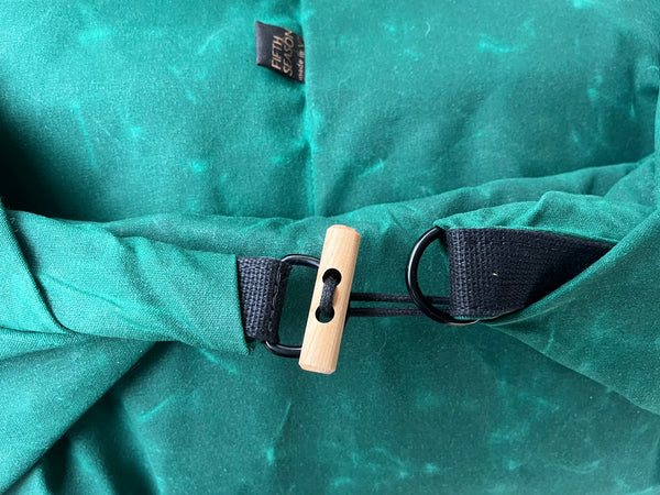 Close-up of a teal-green fabric bag or garment with a black strap and a light natural wood toggle closure, plus a small black fabric tag near the top.