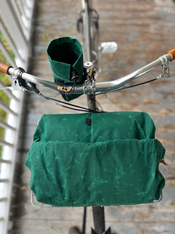 A green bicycle basket cover and matching green handlebar bag are shown mounted on a bicycle over a wooden bridge.