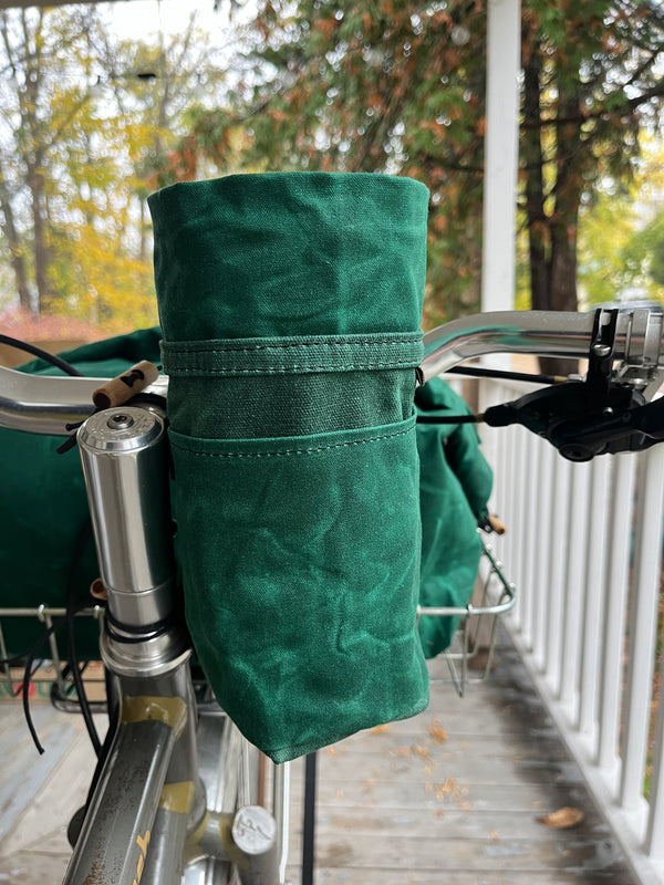 A dark green fabric handlebar bag mounted on a bicycle handlebar, with an outdoor porch and trees in the background.