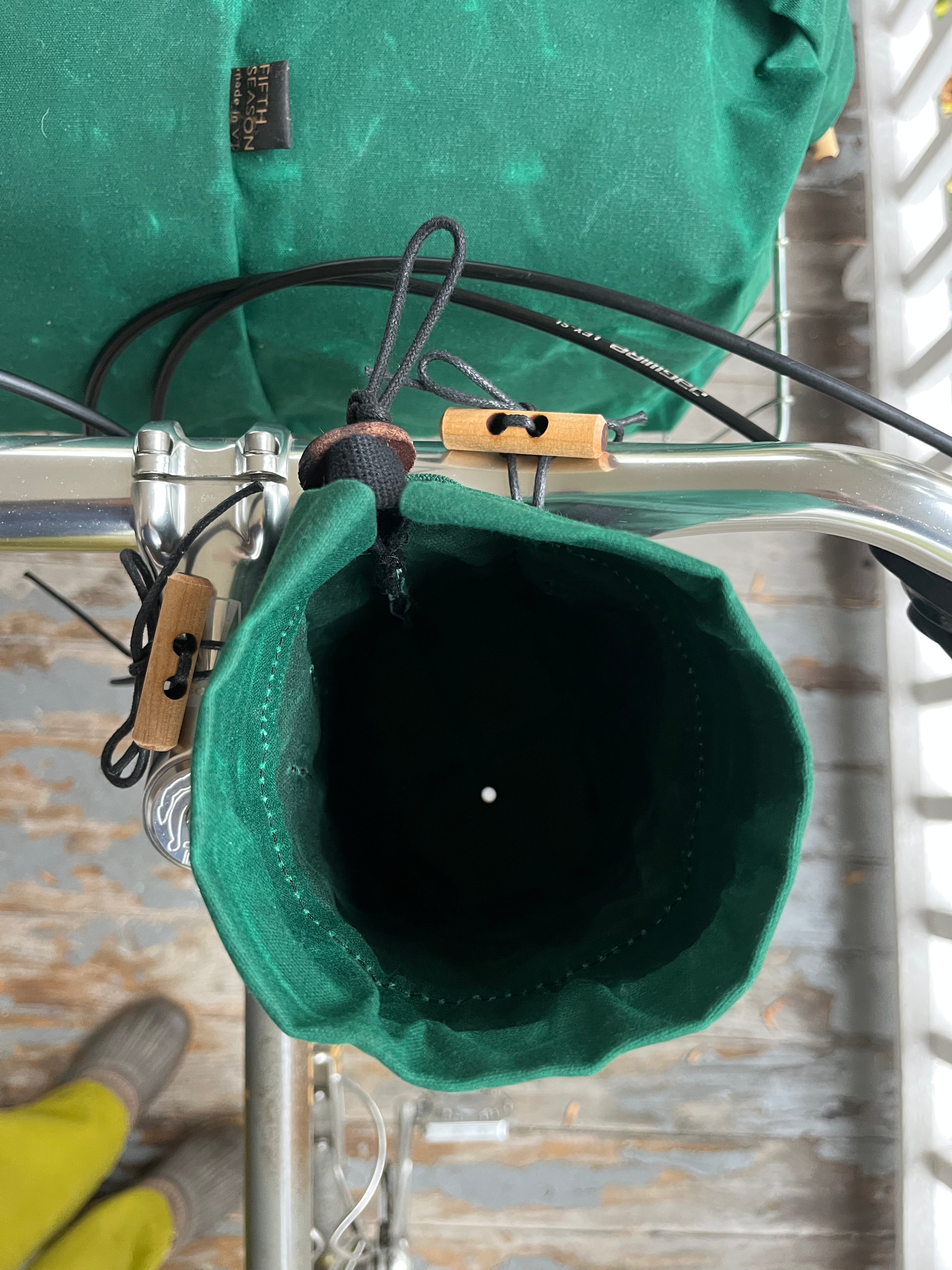 A close-up of a green fabric bike bag hanging from a bicycle frame, with a second green bag mounted above it. The bags are attached with black straps and tan plastic clips, shown against a rustic wooden background.