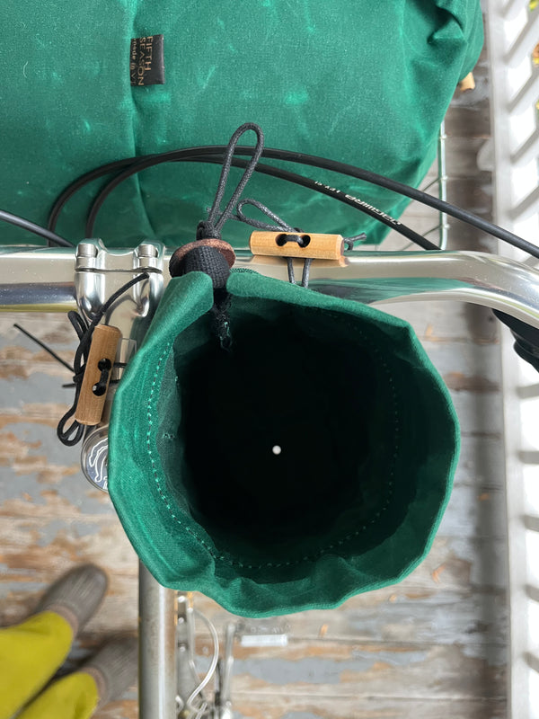 A close-up of a green fabric bike bag hanging from a bicycle frame, with a second green bag mounted above it. The bags are attached with black straps and tan plastic clips, shown against a rustic wooden background.