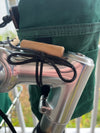 A close-up of a green fabric bag or backpack strap attached to a shiny silver metal handlebar with black cord ties and a small wooden block, photographed outdoors with blurred railings and grass in the background.