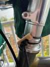 Close-up of a bicycle headset area with a silver metal stem, dark green fabric cover, black cable, and a small tan wooden stopper or cap tied with a black cord.