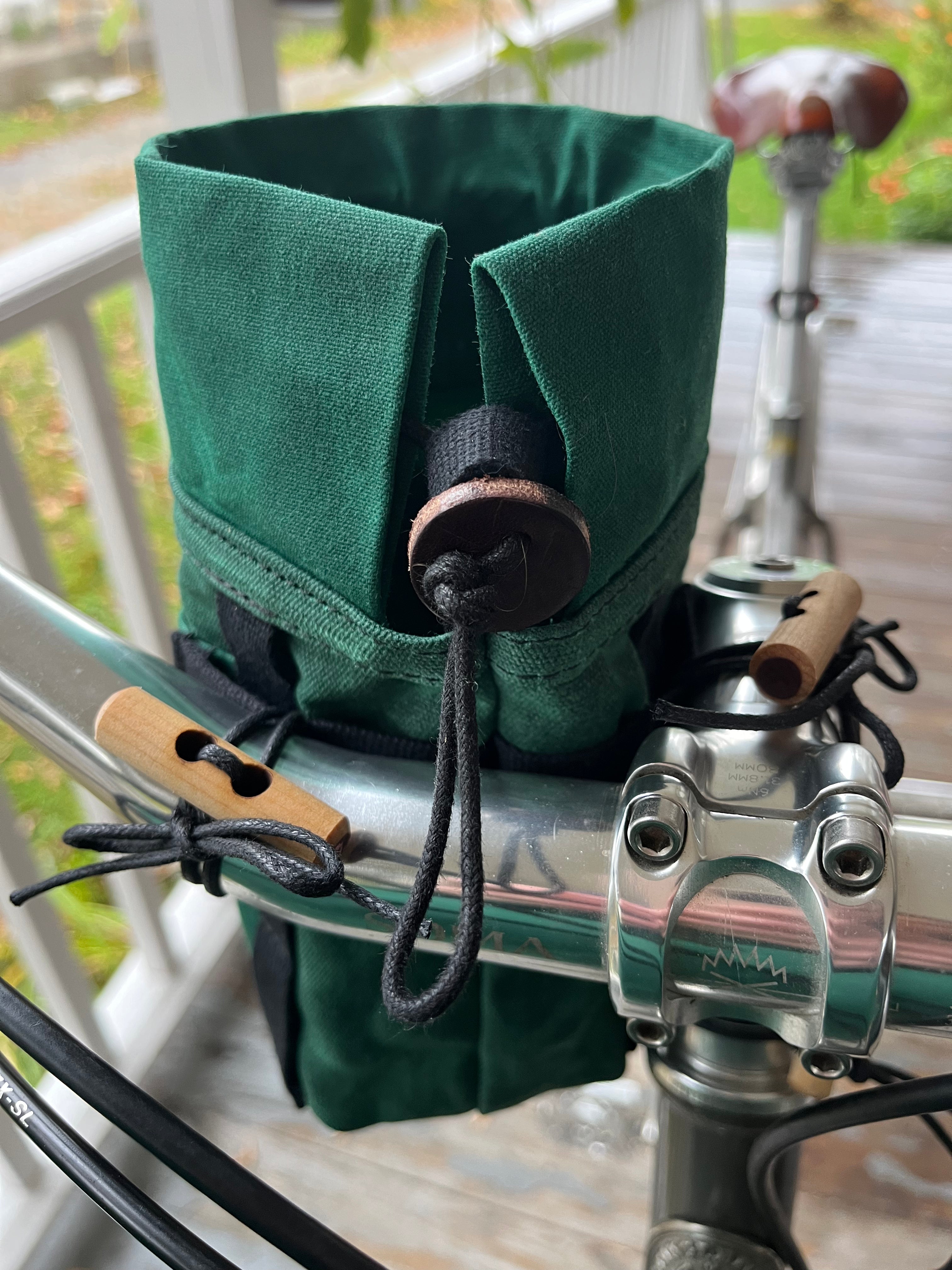 A dark green fabric bicycle handlebar bag with a drawstring closure and wooden toggle, mounted on a bicycle handlebar on a wooden porch.