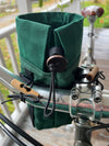 A dark green fabric bicycle handlebar bag with a drawstring closure and wooden toggle, mounted on a bicycle handlebar on a wooden porch.