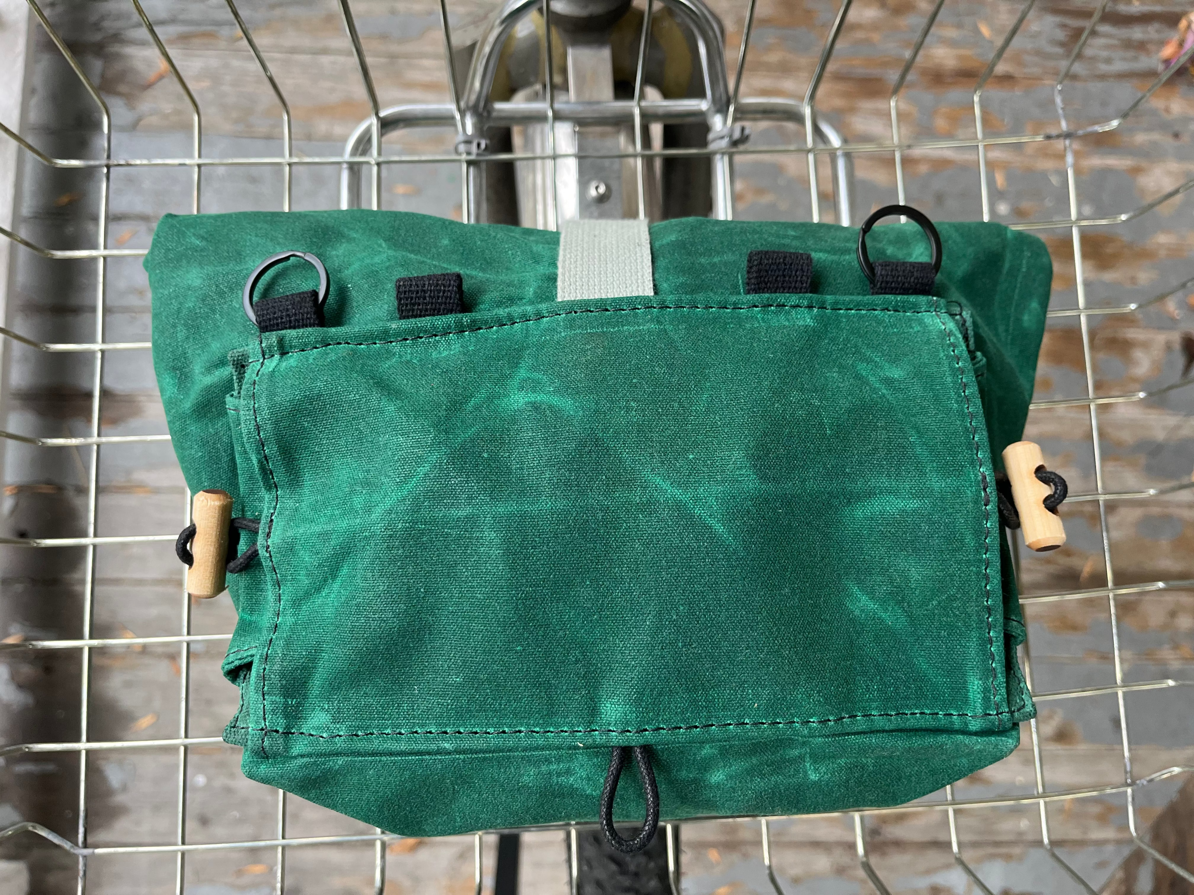 A green waxed canvas bicycle pannier bag hanging from a metal wire cart, showing its front flap, black loops, and wooden toggle hardware.