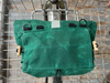 A green waxed canvas bicycle pannier bag hanging from a metal wire cart, showing its front flap, black loops, and wooden toggle hardware.