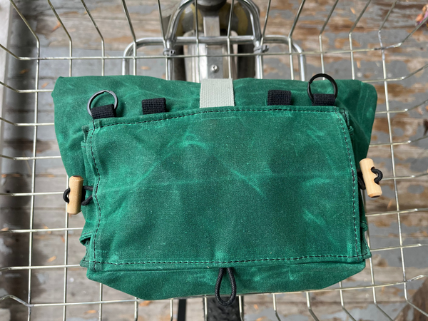 A green waxed canvas bicycle pannier bag hanging from a metal wire cart, showing its front flap, black loops, and wooden toggle hardware.
