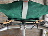 A close-up of a dark green bicycle pannier bag mounted on a silver bicycle frame with black straps and light wood toggle fasteners. A gray webbing strap is visible across the front of the bag.
