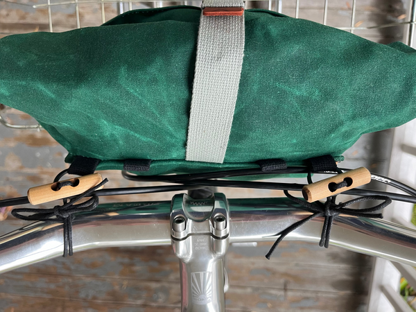 A close-up of a dark green bicycle pannier bag mounted on a silver bicycle frame with black straps and light wood toggle fasteners. A gray webbing strap is visible across the front of the bag.