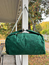 A dark green canvas duffel bag with a matching top handle and shoulder strap, hanging from a white porch post with outdoor trees and autumn foliage in the background.