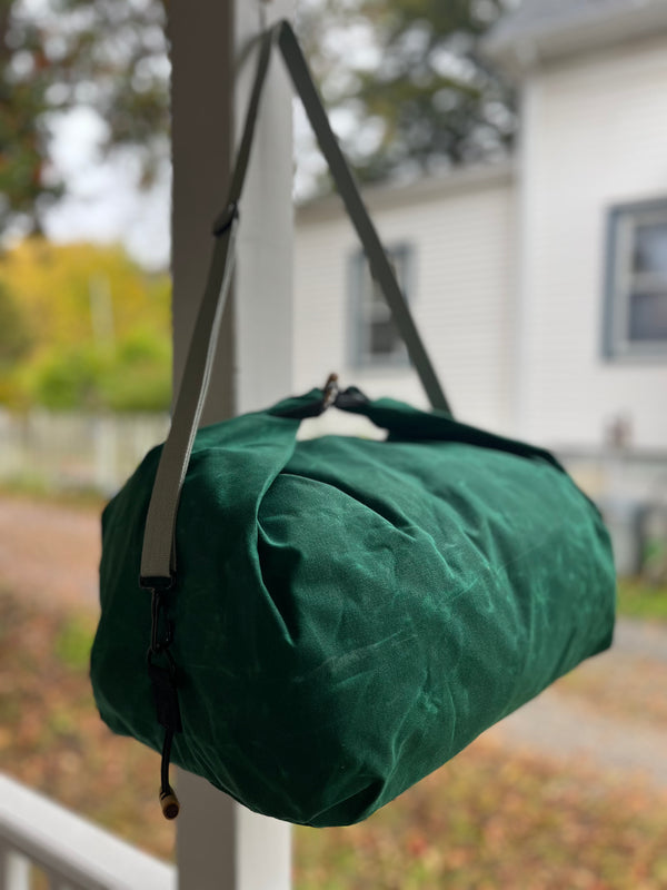 A dark green canvas duffel bag with a gray shoulder strap hanging from a porch post, with a blurred outdoor background showing a house and yard.