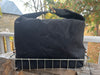 A black canvas duffel bag with a top handle and metal hardware, resting on a wire rack outdoors on a wooden surface with a garden background.