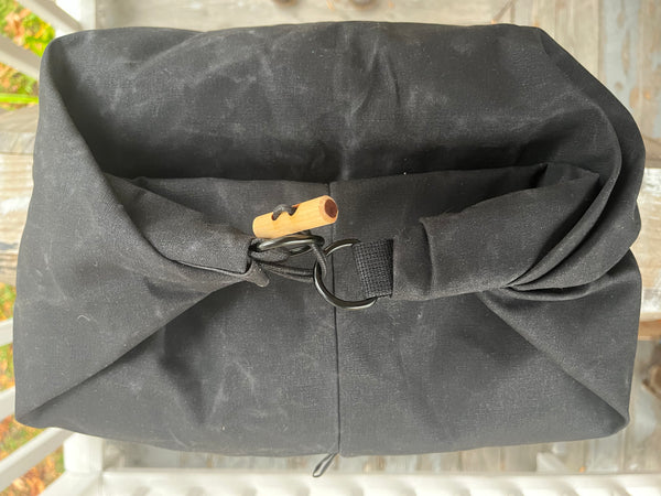 A black canvas dry bag or pouch with a rolled top closure, secured by a black strap and metal ring, featuring a small orange-brown wooden toggle, photographed outdoors on a porch.