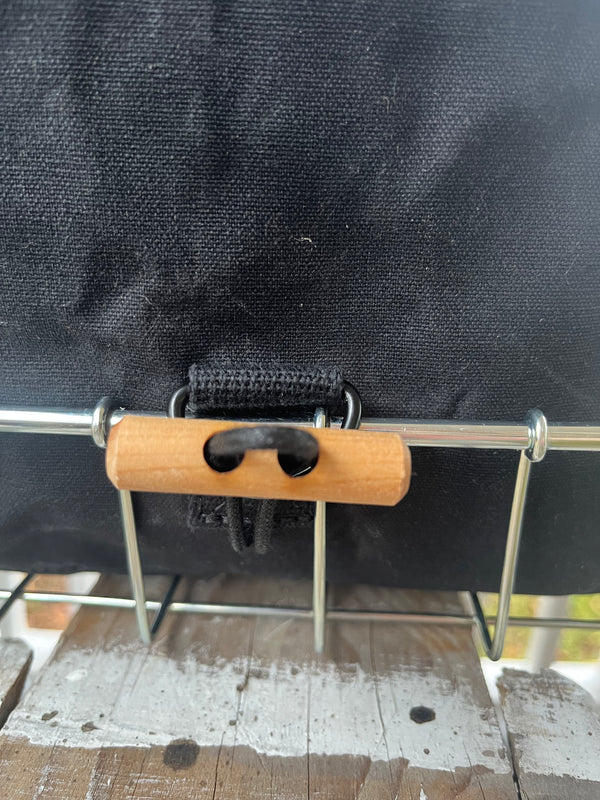 Close-up of a black fabric bag or basket with a metal wire rack and a small wooden handle secured by a black strap.