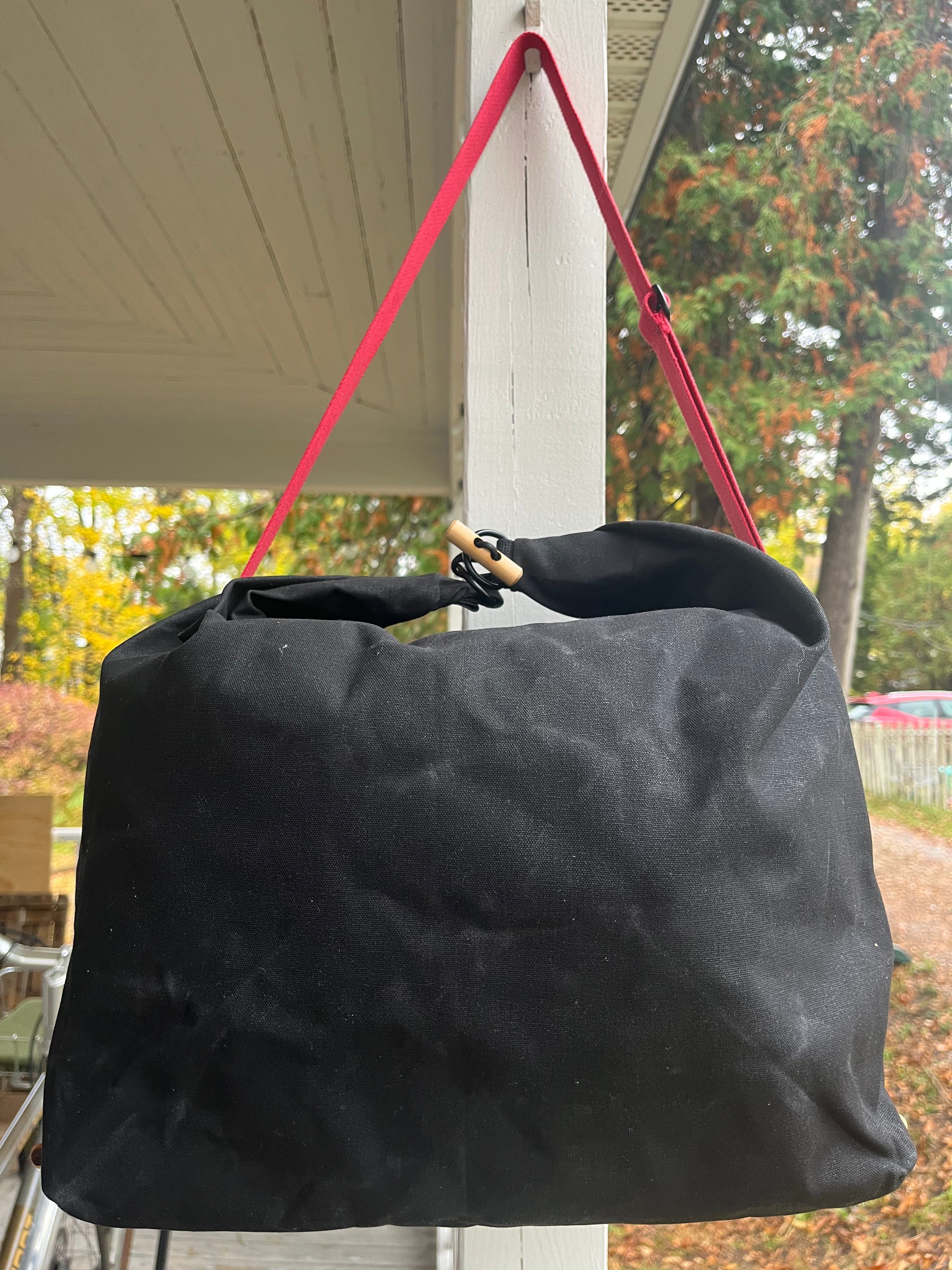 A black canvas tote bag with a bright pink strap hanging from a porch post, with an outdoor yard and trees visible in the background.