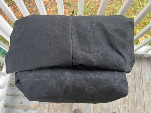 A folded black waxed canvas bag or cover resting on a wooden deck railing with outdoor foliage visible in the background.