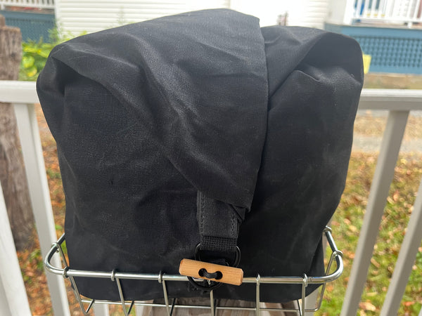 A black fabric bag with a wooden toggle clasp resting on a metal wire bicycle basket, set against an outdoor residential background.