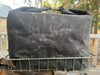 A dark gray waxed canvas tote bag with a top handle, resting on a silver metal wire basket outdoors on a wooden deck with trees in the background.