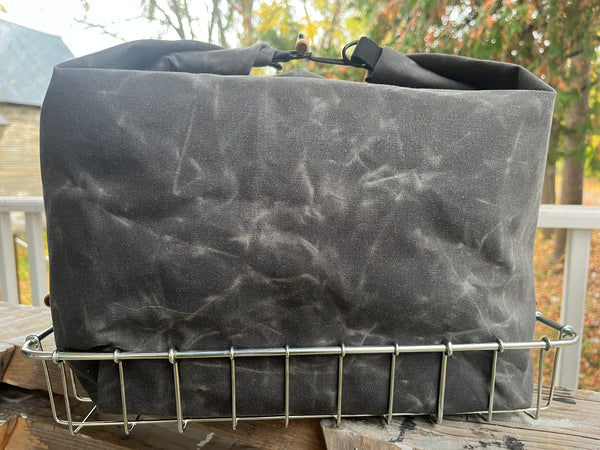 A dark gray waxed canvas tote bag with a top handle, resting on a silver metal wire basket outdoors on a wooden deck with trees in the background.