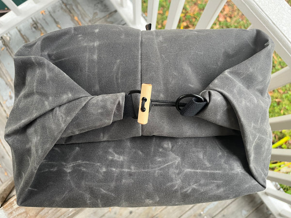 A dark gray waxed canvas bag with a rolled top closure, secured by a black strap and toggle clasp with a light wood accent, photographed outdoors on a wooden deck.