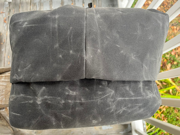 A folded dark gray fabric item, likely a blanket or throw, with a distressed waxed-paper texture, resting on a white porch chair outdoors.