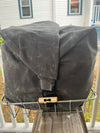 A dark gray waxed canvas backpack or gear bag with a roll-top closure and a small wooden accent, resting on a metal bike rack on a porch with a house in the background.