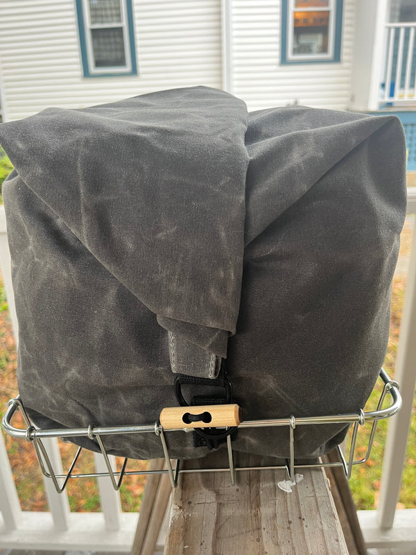 A dark gray waxed canvas backpack or gear bag with a roll-top closure and a small wooden accent, resting on a metal bike rack on a porch with a house in the background.