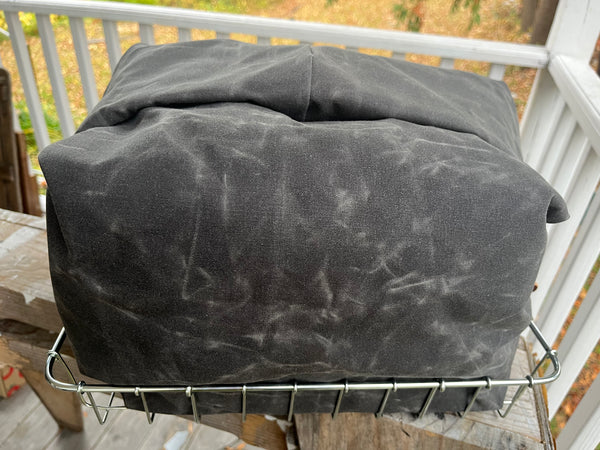 A dark gray waxed canvas cover, likely for a grill or smoker, draped over a metal wire rack on a wooden deck with a white railing and outdoor foliage in the background.