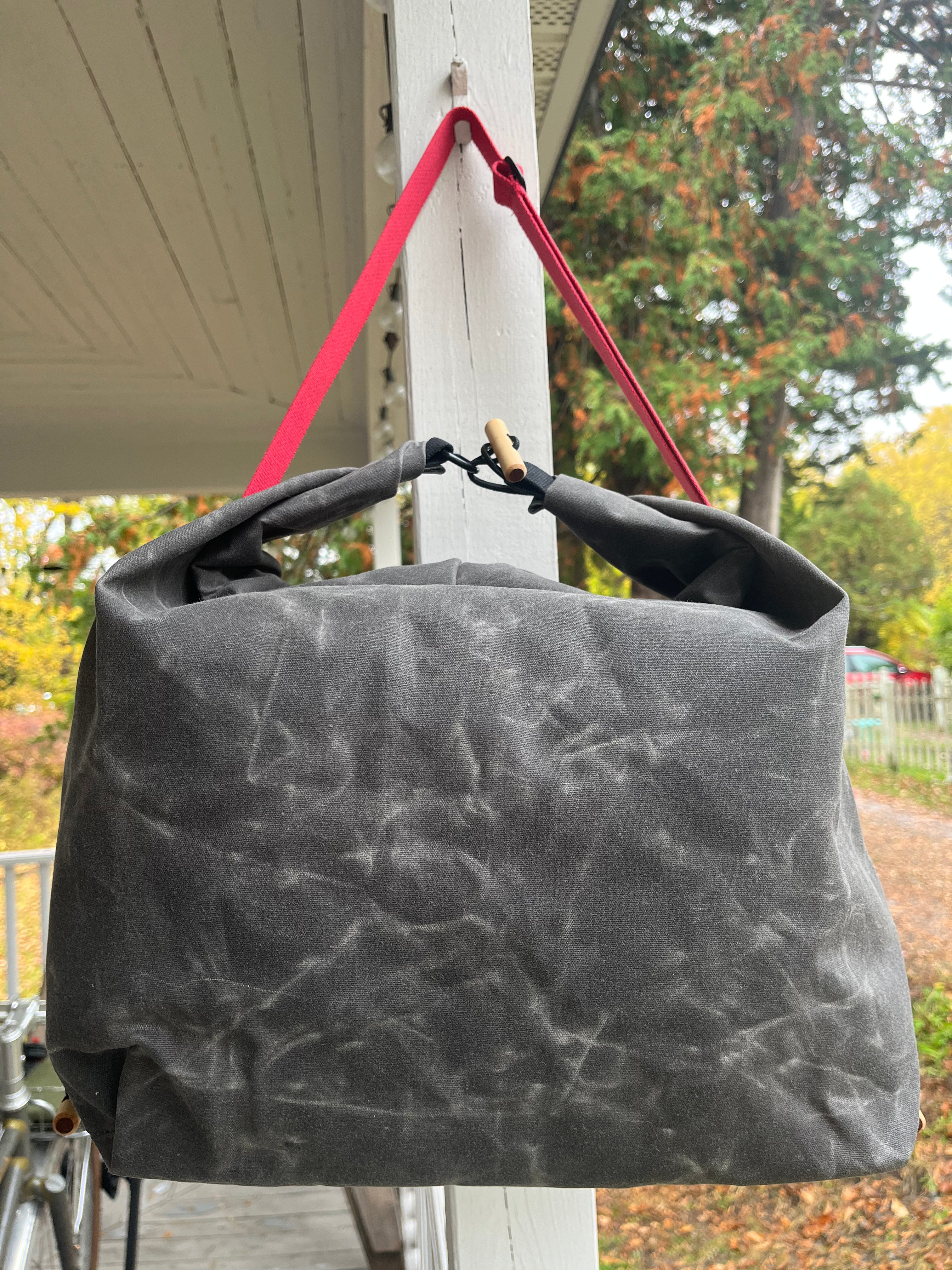 A dark gray waxed canvas tote bag with a bright pink strap hanging from a white porch post, with an outdoor garden background.