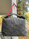 A dark gray waxed canvas tote bag with a bright pink strap hanging from a white porch post, with an outdoor garden background.