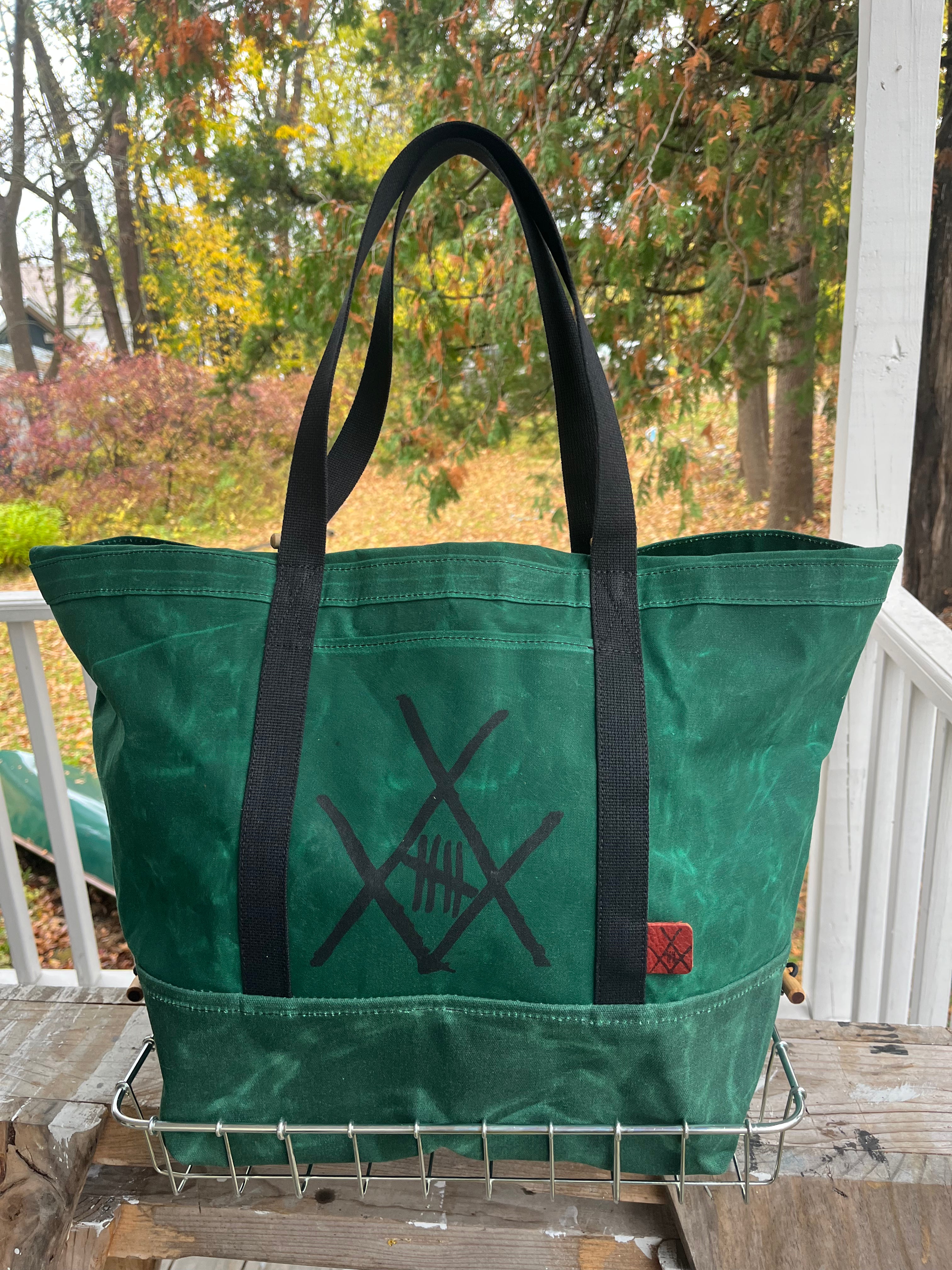 A green canvas tote bag with black handles and a black crossed-knife style graphic on the front, sitting on a metal wire rack on a porch with an outdoor autumn garden background.