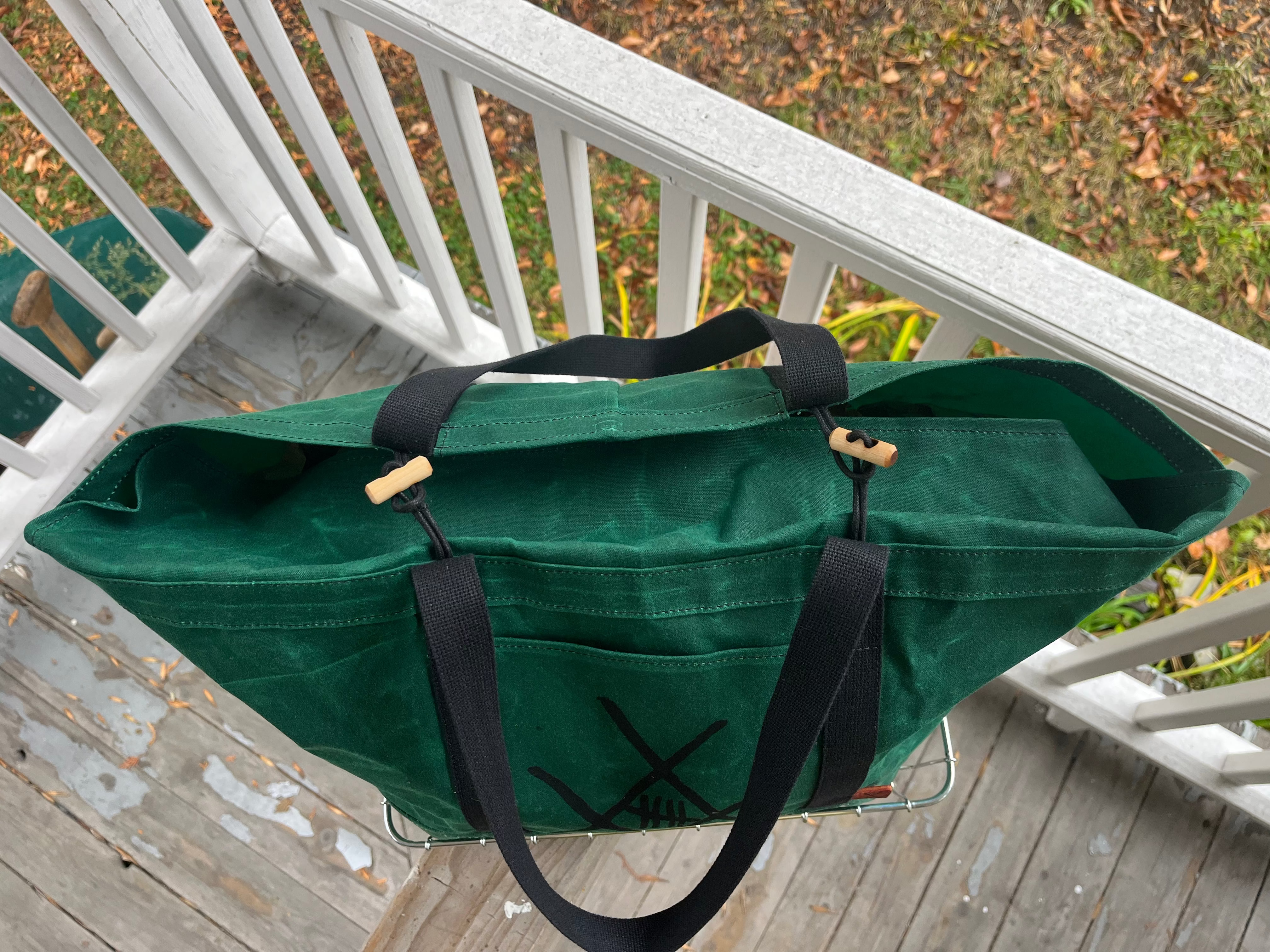 A dark green canvas tote bag with black straps and wooden toggle closures, photographed outdoors on a white porch railing with a wooden deck and grass visible.
